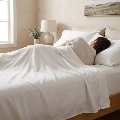 A woman asleep under the white sheet set in a softly lit bedroom, showing the flat sheet turned back over the quilt